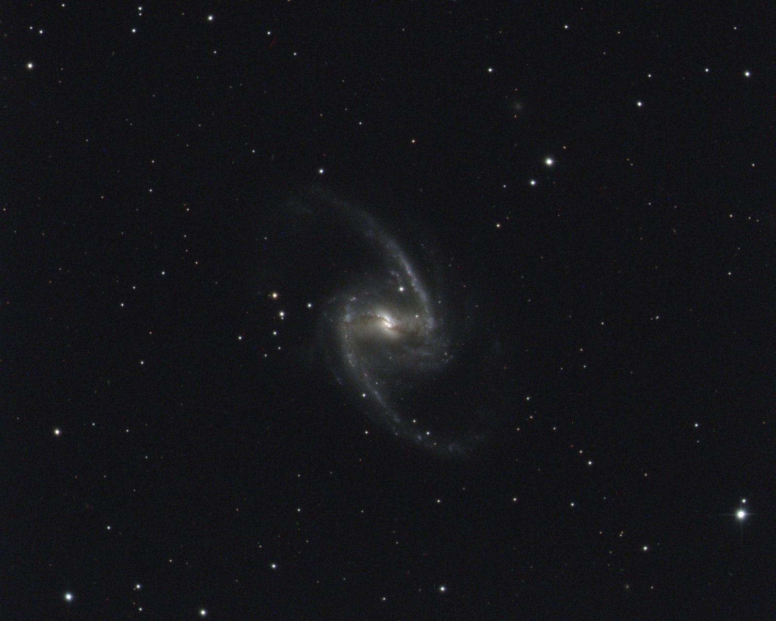 A barred spiral galaxy seen nearly face on: a bright core with a straight bar of stars through it, a dark dust lane across the nucleus, and two long arms curling off the ends of the bar into an S, dotted with blue knots, against a dark field of stars.