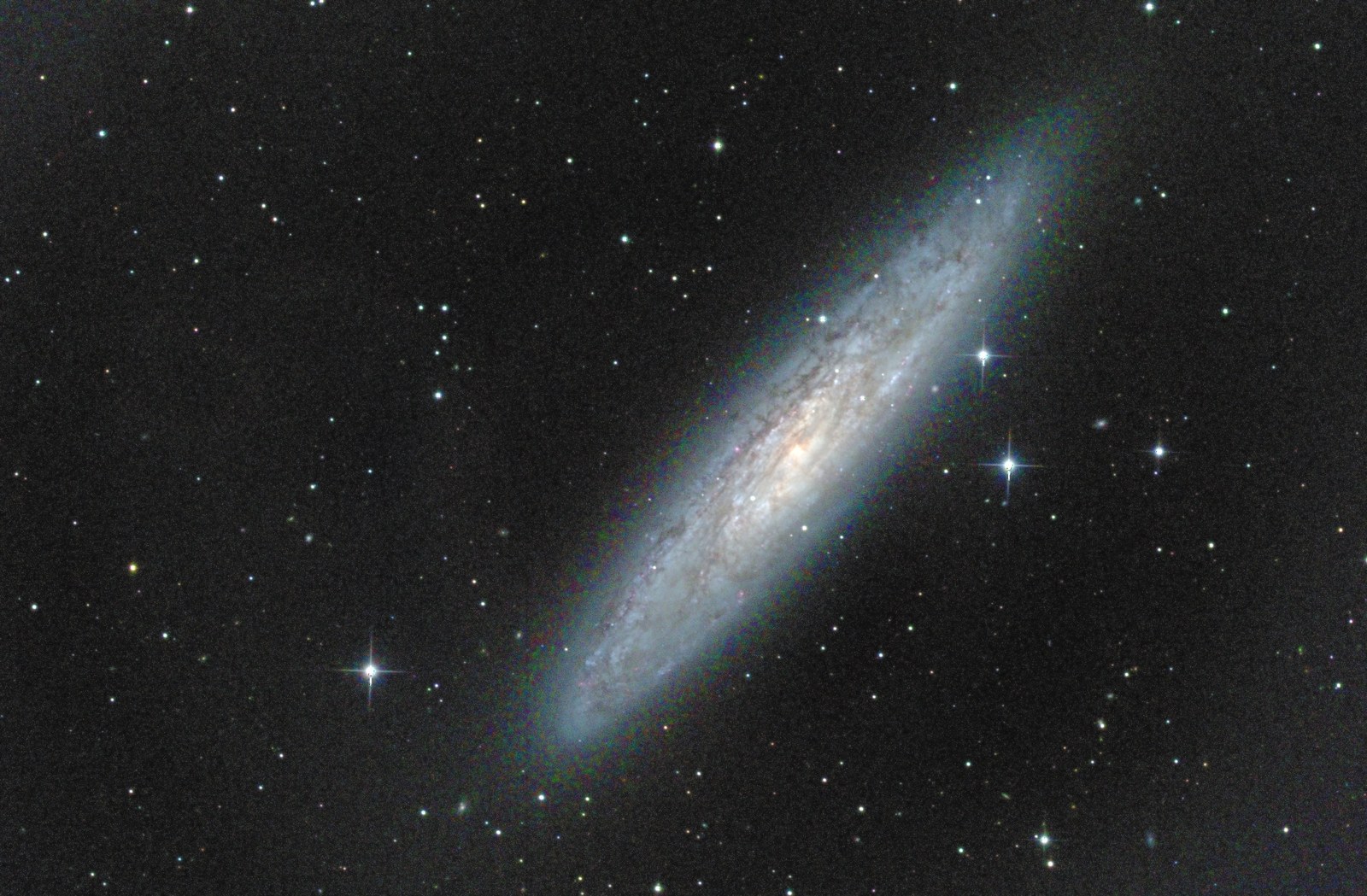 A long spiral galaxy seen almost edge-on, tilted from lower left to upper right across a dark star field, its disk streaked with dark dust lanes, a warm gold glow at the center and a pale blue haze along the arms.