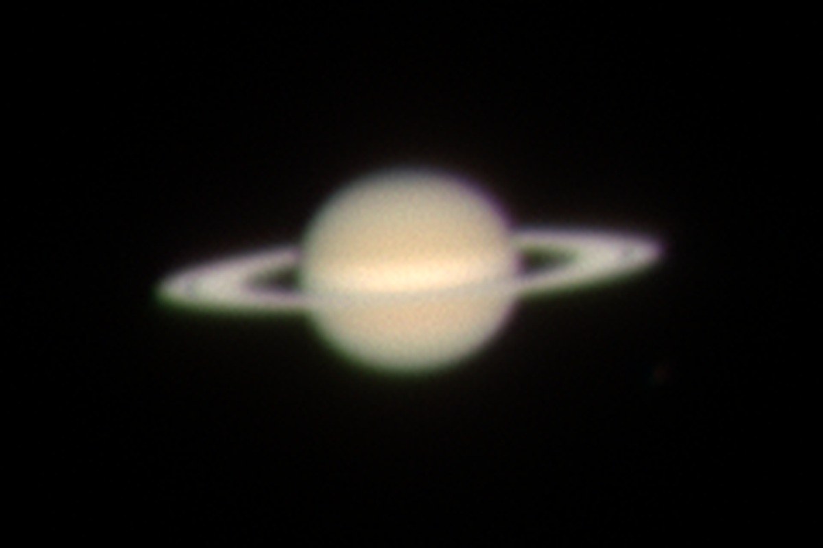 Saturn against black sky, a pale gold globe with a brighter band across its middle, crossed by the rings seen almost edge-on as a thin bright blade, with a dark gap between ring and planet on each side.