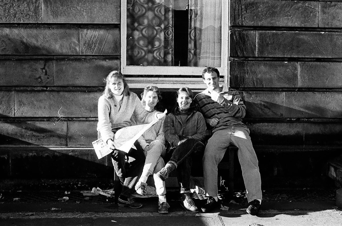 Four students on a bench against a sandstone wall in low sun, a newspaper open on a lap.