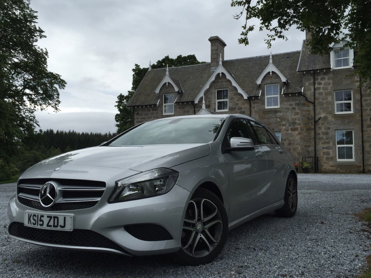 A silver Mercedes hatchback on gravel in front of a stone Victorian house with pointed dormers.