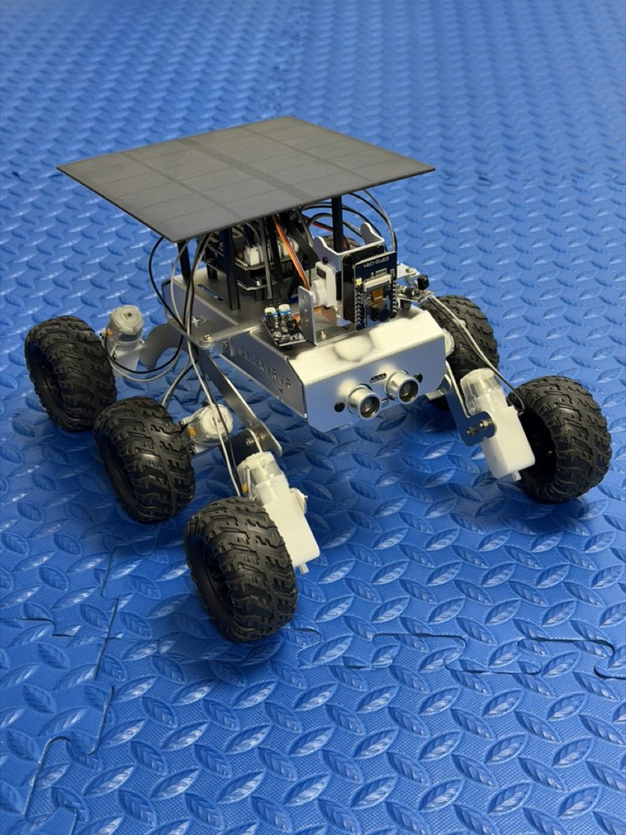 A small six-wheeled silver rover with a square solar panel on top and two round ultrasonic sensors in front, on a blue foam mat.